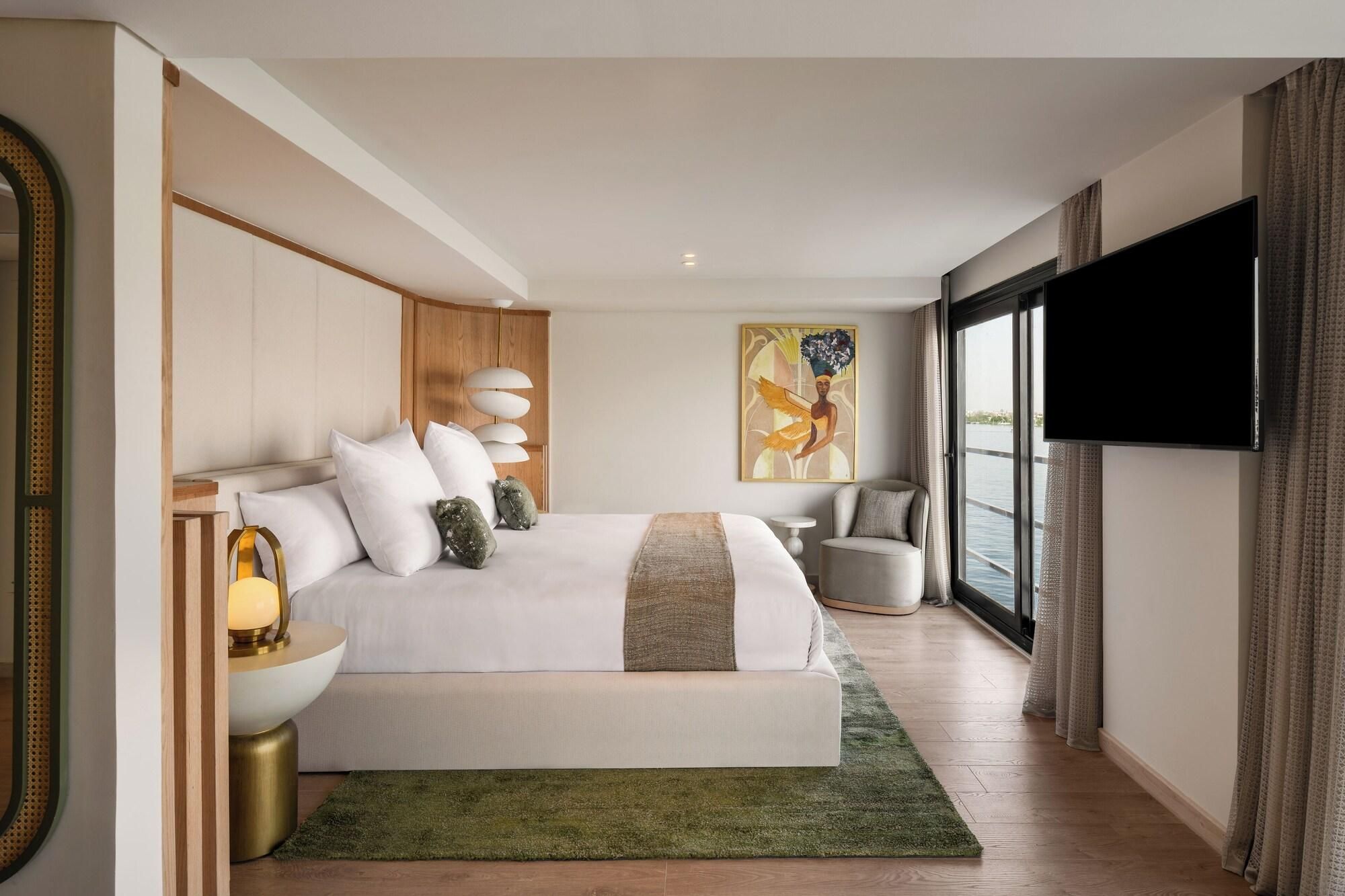 A modern hotel room with a large bed, a flat-screen TV, a painting, and a view of the water.