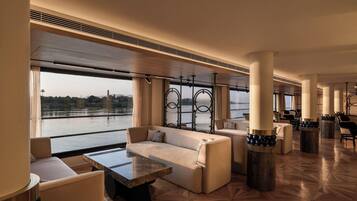 A spacious lounge area with a large window offering a view of a body of water, a marble coffee table, and comfortable seating.