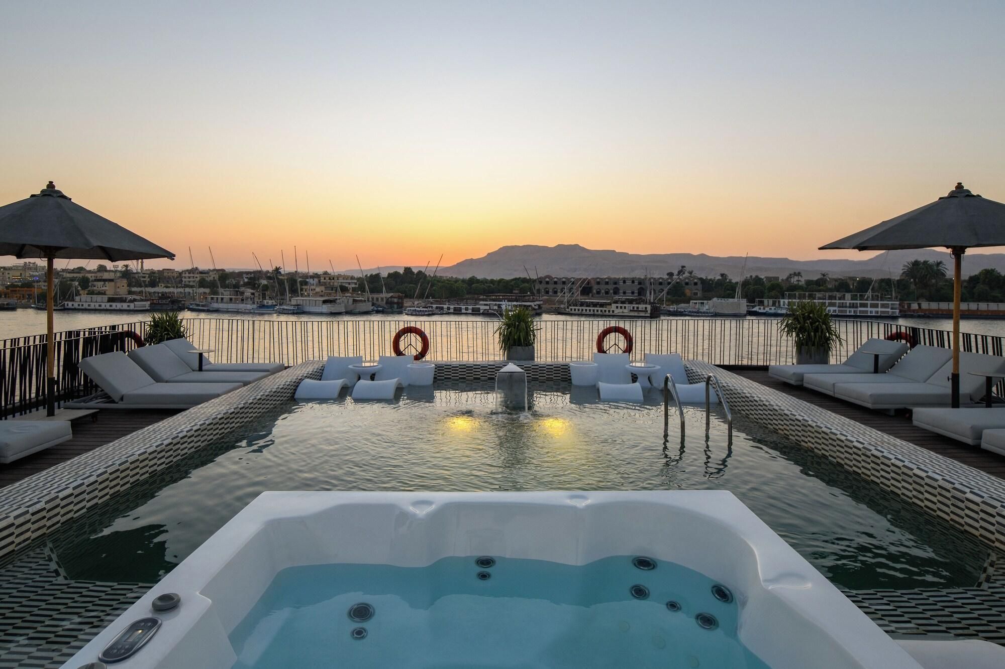A rooftop pool with lounge chairs, a hot tub, and a view of the sunset over a marina.