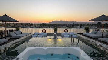 A rooftop pool with lounge chairs, a hot tub, and a view of the sunset over a marina.