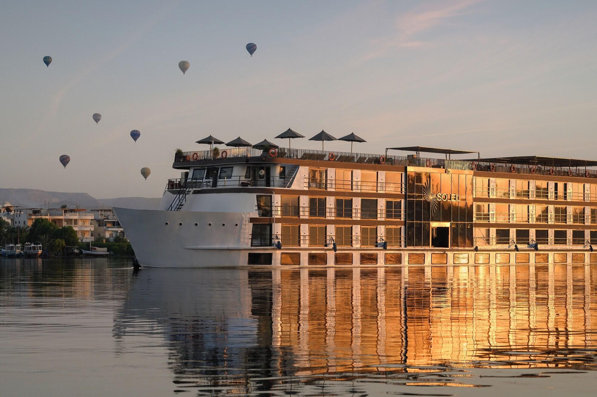 A large riverboat with multiple decks, outdoor seating with umbrellas, and a reflection on the water.