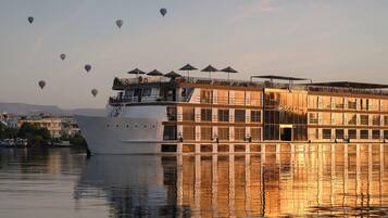 A large riverboat with multiple decks, outdoor seating with umbrellas, and a reflection on the water.