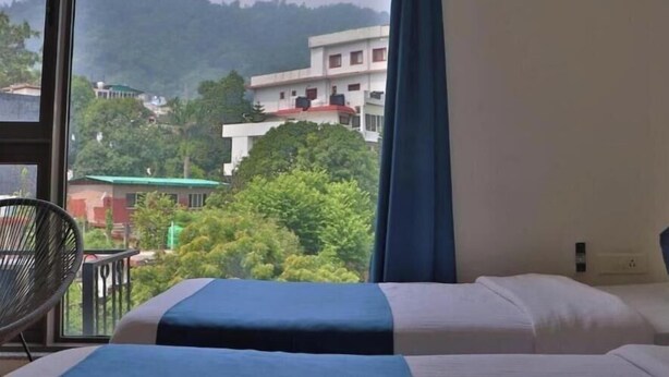 A hotel room with a bed, a chair, and a view of buildings and trees. - The Blue Jay Hostel Rishikesh (Narendranagar)