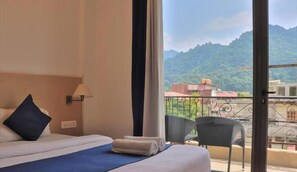 A hotel room with a bed, a chair, a window with a view of mountains and buildings. - The Blue Jay Hostel Rishikesh (Narendranagar)