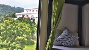 A room with a bed, a window with a view of a building and trees, and a green curtain. - The Blue Jay Hostel Rishikesh (Narendranagar)