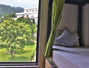 A room with a bed, a window with a view of a building and trees, and a green curtain. - The Blue Jay Hostel Rishikesh (Narendranagar)