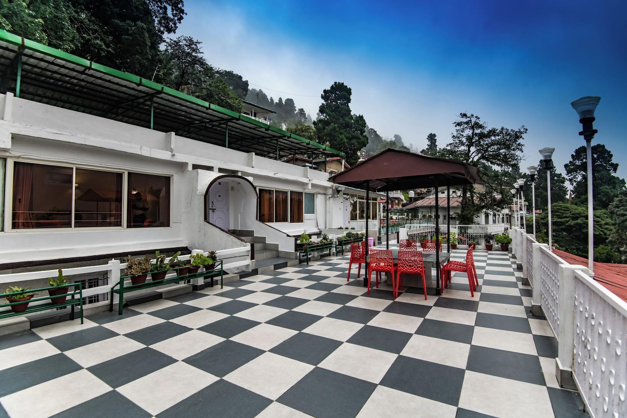 A checkered outdoor patio with red chairs and tables, a covered seating area, and a white building with large windows and a green roof.