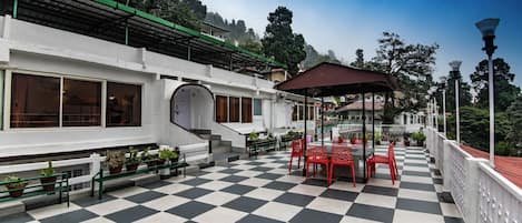 A checkered outdoor patio with red chairs and tables, a covered seating area, and a white building with large windows and a green roof.