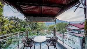 A balcony with a table and chairs, a view of trees and mountains, and a building with a red roof in the background.