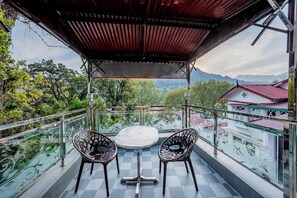 A balcony with a table and chairs, a view of trees and mountains, and a building with a red roof in the background.