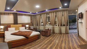 A modern hotel room with a large bed, a small table, two armchairs, a balcony with a railing, and a TV mounted on the wall.