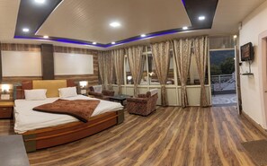 A modern hotel room with a large bed, a small table, two armchairs, a balcony with a railing, and a TV mounted on the wall.