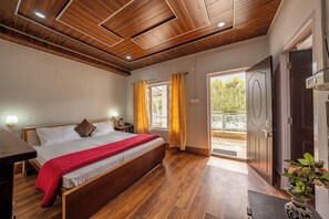 A bedroom with a wooden ceiling, a bed with a red blanket, a bedside table, a lamp, and a view of the outdoors through a window with yellow curtains. - India Hotel (Nainital)