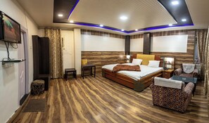 A hotel room with a bed, bedside tables, a chair, a TV, and a wooden floor. - India Hotel (Nainital)
