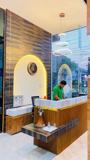 A hotel reception desk with a marble counter, a wooden panel design, a clock, and a modern light fixture. - Hotel Aranyasutra Inaya Indore (Indore)