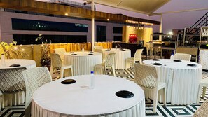 An outdoor dining area with round tables and chairs, a stage with musical equipment, and a view of a building in the background. - Hotel Aranyasutra Inaya Indore (Indore)
