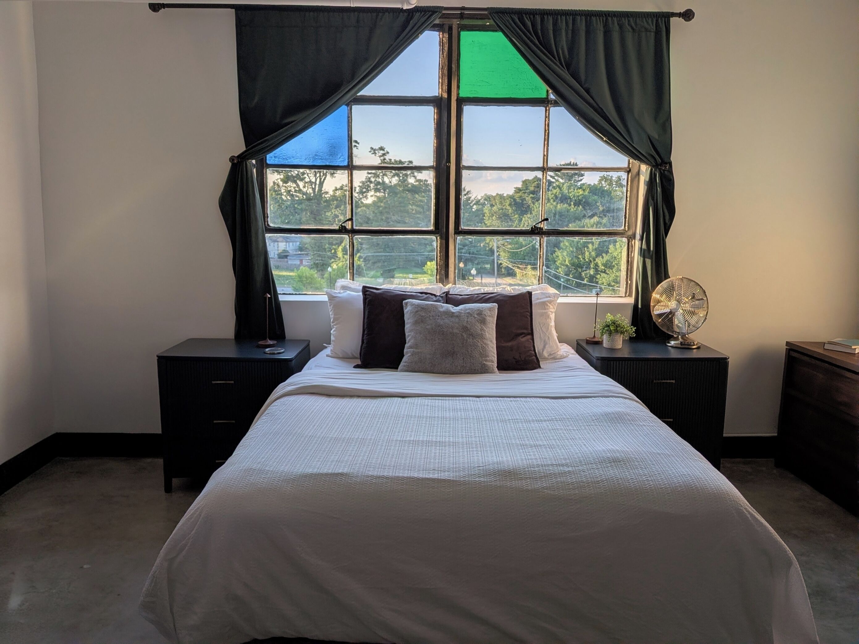 A bedroom with a large window, a bed with pillows, a bedside table with a lamp, and a nightstand with a fan.