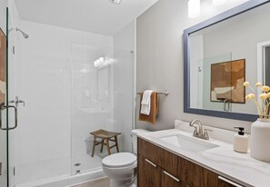 A modern bathroom with a glass shower enclosure, a wooden stool, a white toilet, and a vanity with a mirror.