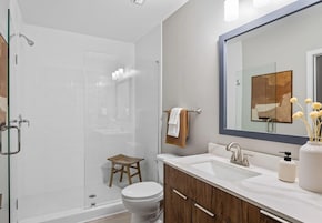 A modern bathroom with a glass shower enclosure, a wooden stool, a white toilet, and a vanity with a mirror.