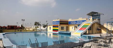 Zona de piscina con sillones y mesas, un tobogán y un edificio con fachada colorida.