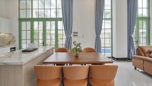 Een eetruimte met een houten tafel en zes stoelen, een keuken met marmeren aanrechtbladen en grote ramen met groene kaders en blauwe gordijnen.