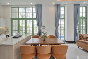 Ein Essbereich mit einem Holztisch und sechs Stühlen, eine Küche mit Marmorarbeitsplatten und große Fenster mit grünen Rahmen und blauen Vorhängen.