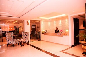 Ein Hotel-Empfangsbereich mit Empfangstresen, einer Wartezone mit Sitzgelegenheiten und einem Essbereich im Hintergrund.