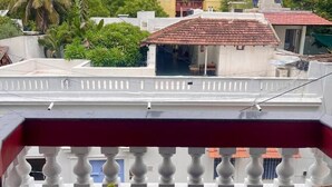 A balcony with a wicker chair and a small table, overlooking a residential area with buildings and greenery. - Le Clarisse Guest House (Chennai)