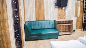 Una habitación con sofá verde, un mueble de madera, televisor montado en la pared y cama blanca.
