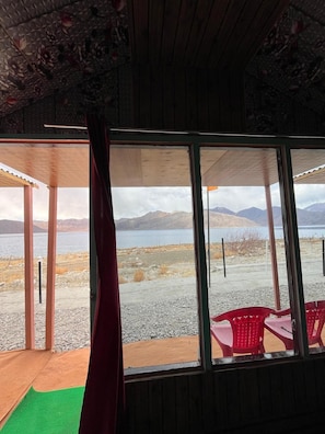 A room with a large window overlooking a lake and mountains, wooden walls, and a green mat on the floor. - Padma Cottage Pangong (Leh)