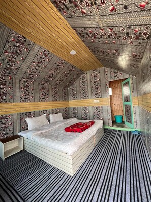 A room with a sloped ceiling, a bed with a red blanket, a wooden headboard, and a small green door. - Padma Cottage Pangong (Leh)
