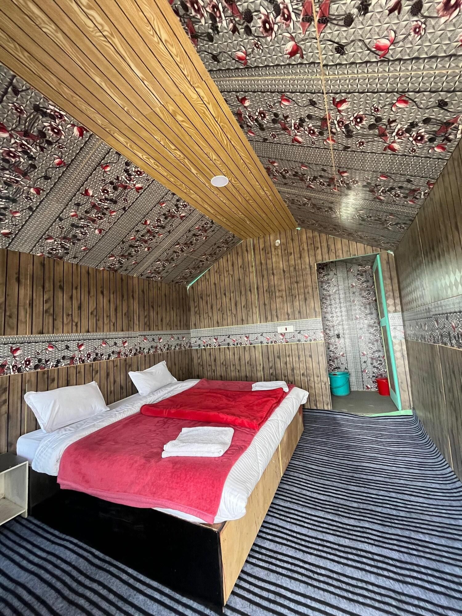 A room with a wooden ceiling, a bed with a red blanket, and a striped rug.