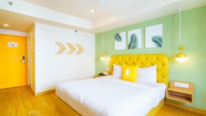Quarto de hotel com uma cama, uma porta de madeira, uma parede com três quadros com padrão de folhas e uma mesa de cabeceira com uma luminária.