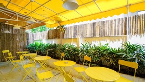Área de estar ao ar livre com mesas e cadeiras amarelas, paredes de bambu e luminárias pendentes.