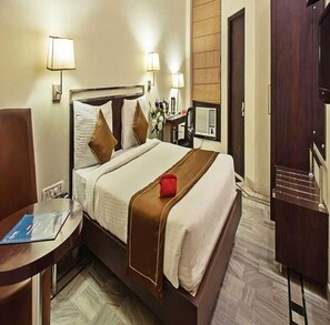 A hotel room with a bed, bedside table, lamp, and a desk with a chair. - Hotel Arch Inn (Jaipur)