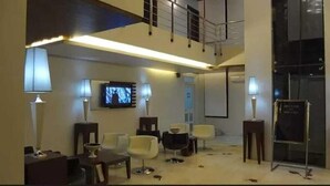 A hotel lobby with a high ceiling, a television mounted on the wall, several chairs, a small table, and a glass partition. - Hotel Arch Inn (Jaipur)