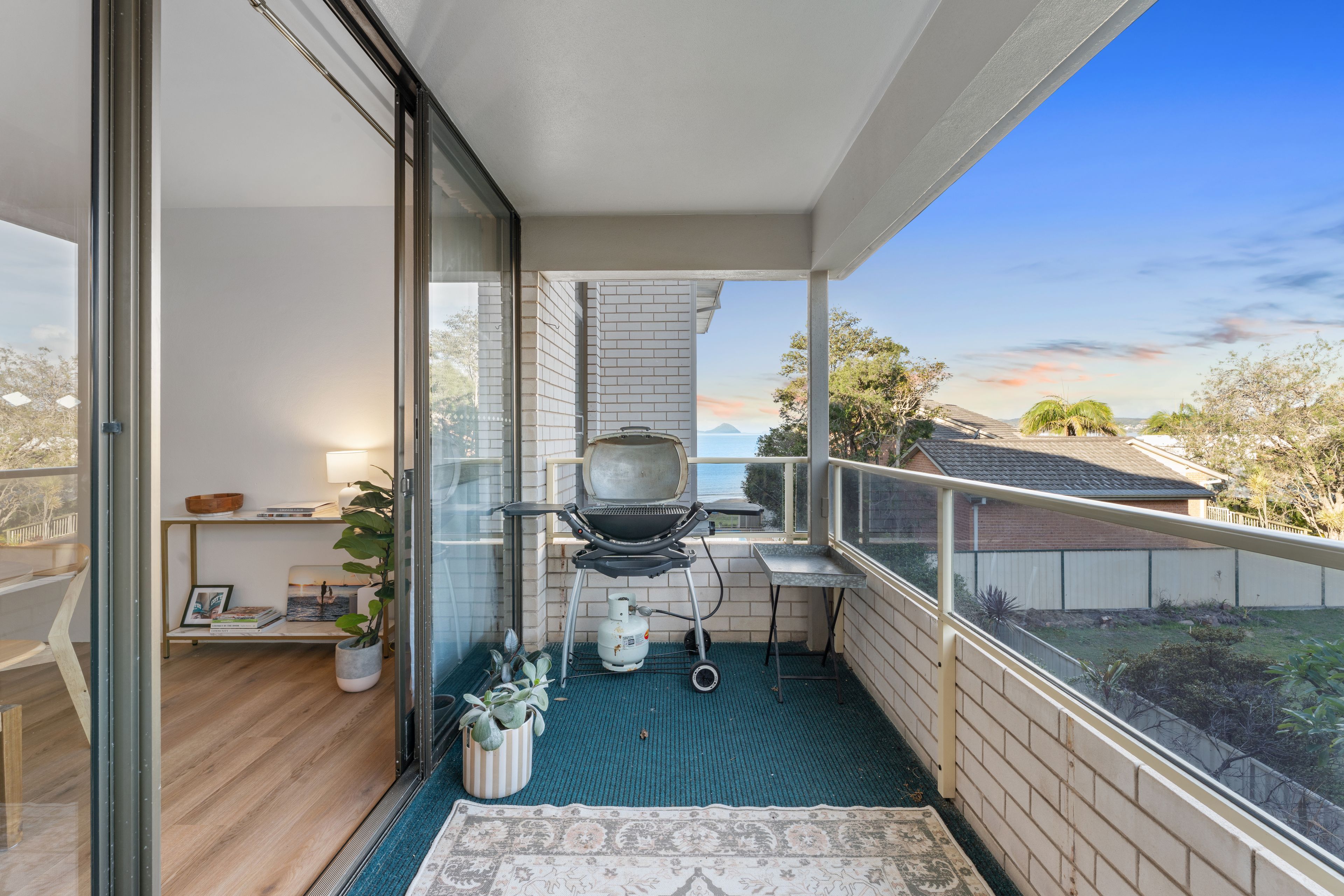 A balcony with a grill, a small table, a chair, a potted plant, and a view of the sea.