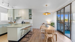 Eine moderne Küche mit einer zentralen Kochinsel, Barhocker, einem Esstisch mit Stühlen und einem großen Schiebeglasfenster zum Balkon.