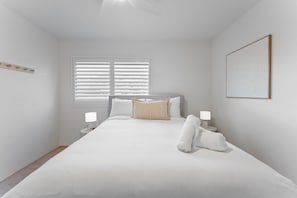 Ein schlicht eingerichtetes Schlafzimmer mit einem Bett, zwei Nachttischlampen, einem Fenster mit Jalousien und einem gerahmten Kunstwerk an der Wand.