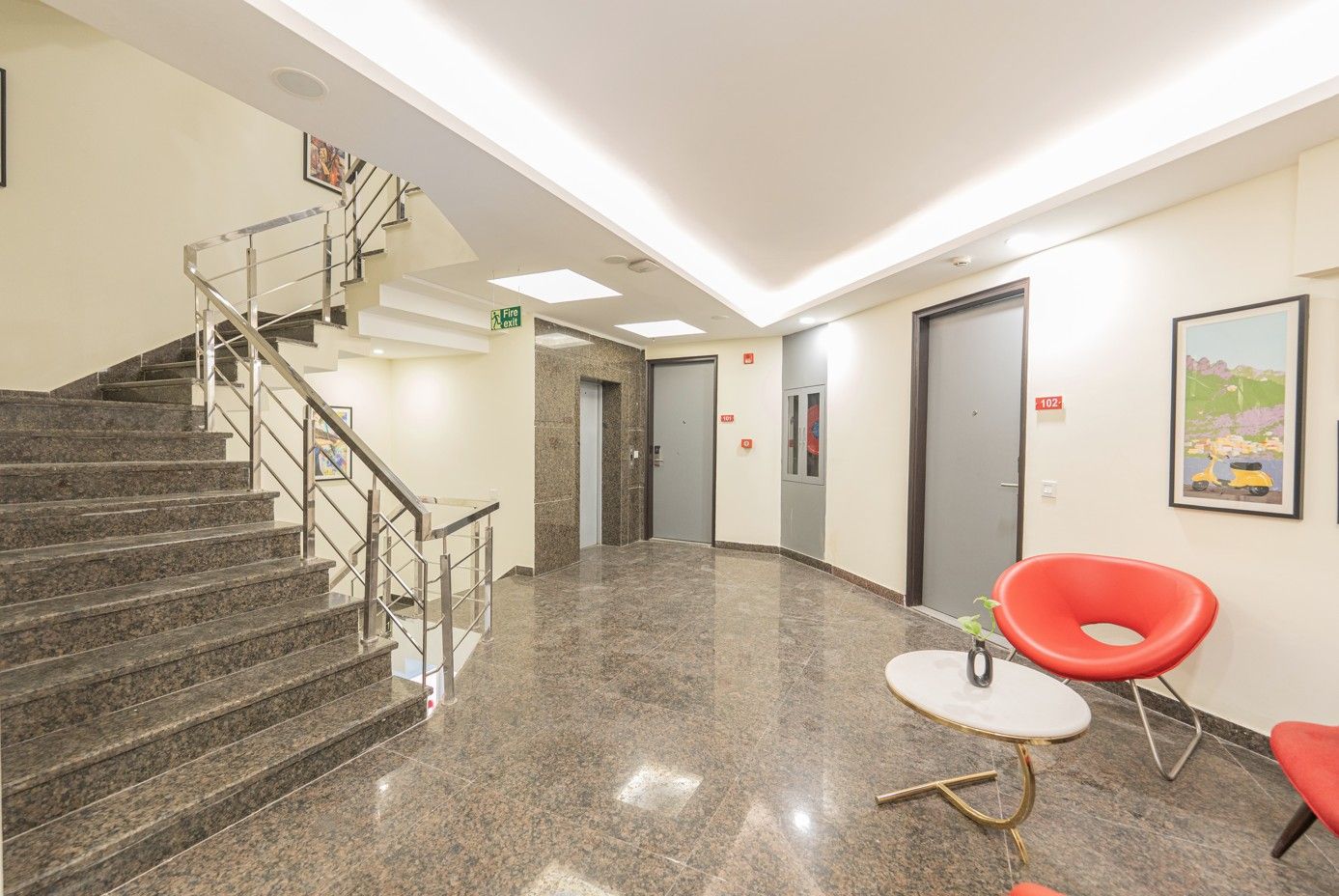 A lobby with a staircase, a fire exit sign, a reception desk, and a door marked with the number 102.