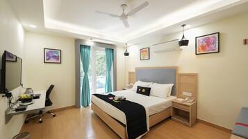 A hotel room with a bed, desk, chair, desk lamp, ceiling fan, artwork, and a window with curtains.