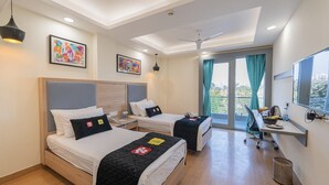 Habitación de hotel con dos camas, un escritorio, una silla, un ventilador de techo y un ventanal grande con cortinas.