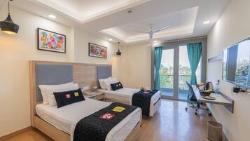 Een hotelkamer met twee bedden, een bureau, een stoel, een plafondventilator en een groot raam met gordijnen.