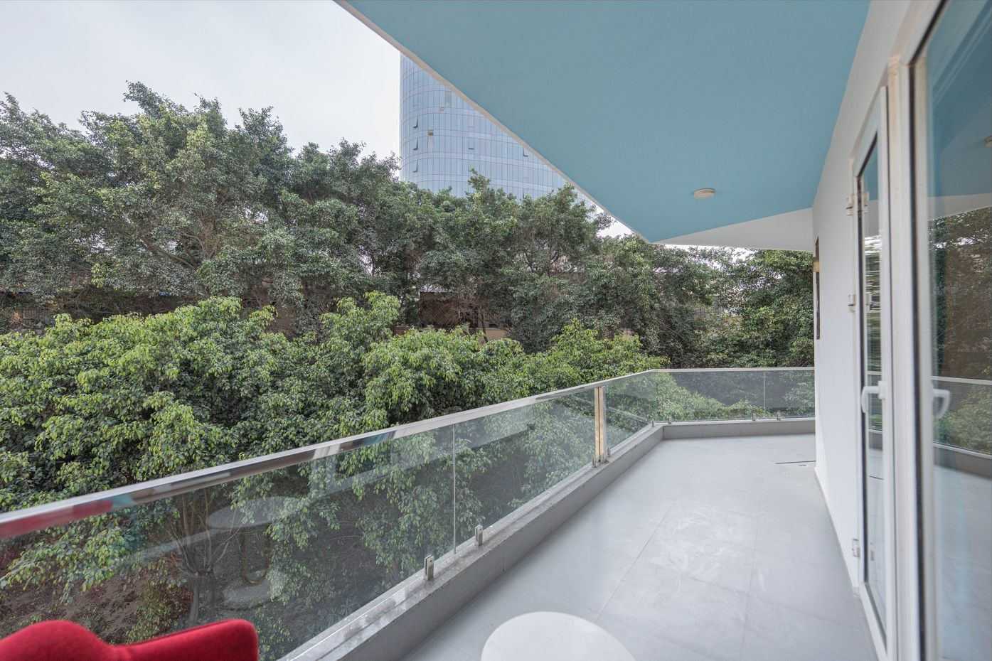 A balcony with a glass railing, a red chair, and a view of dense greenery and a tall building in the background.