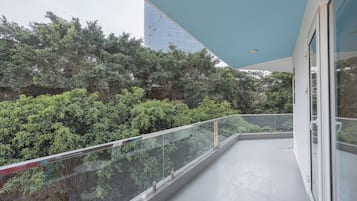 A balcony with a glass railing, a red chair, and a view of dense greenery and a tall building in the background.