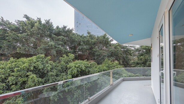 Un balcón con barandilla de cristal, una silla roja y vistas a vegetación densa y un edificio alto al fondo.