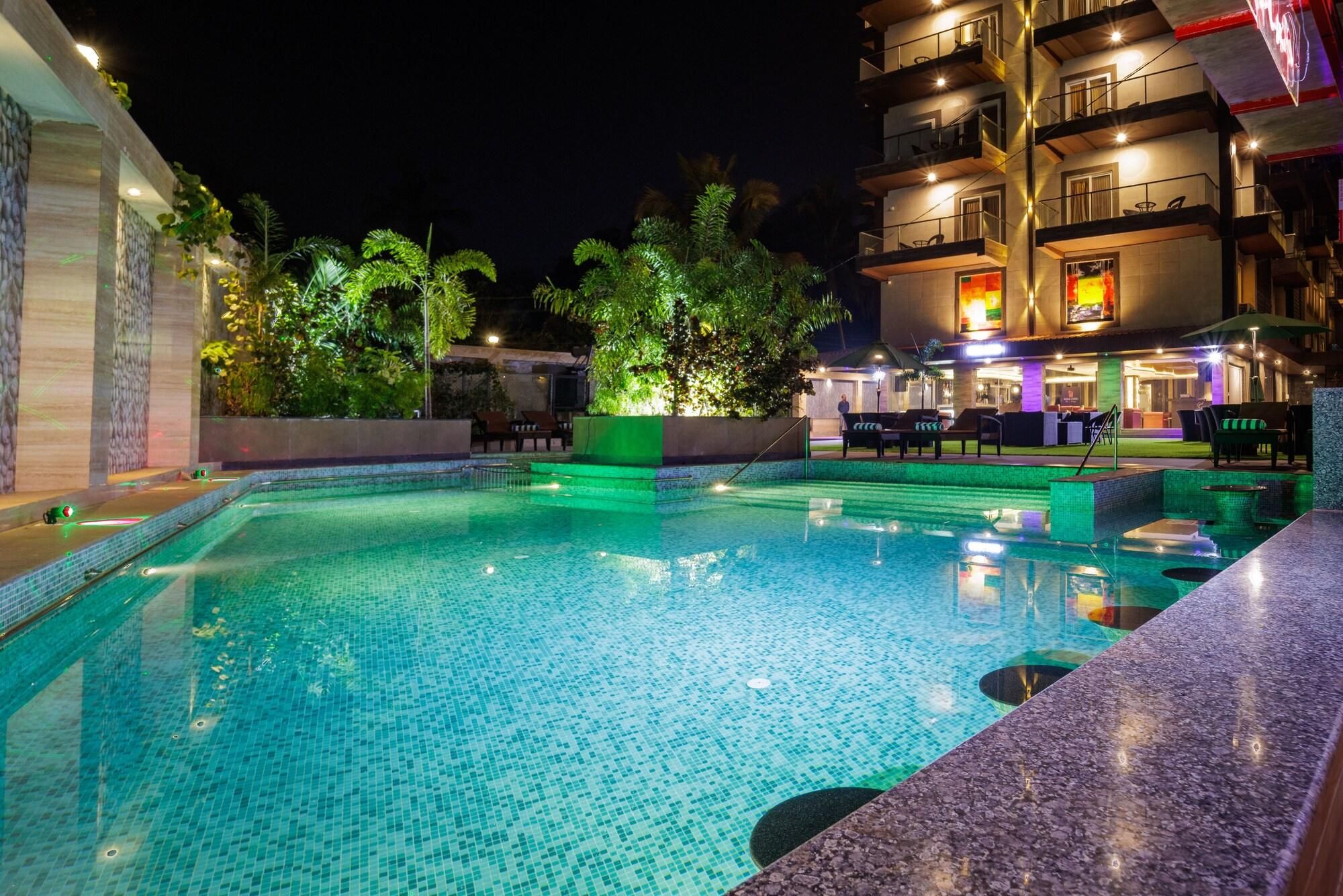 A nighttime swimming pool with tiled flooring, surrounded by greenery and a building with balconies.