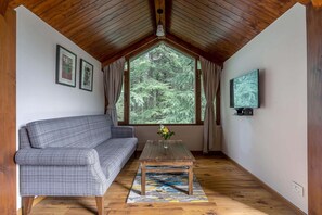 Une pièce chaleureuse avec un plafond en bois, un canapé, une table ornée d’un vase, un téléviseur et une grande fenêtre donnant sur la forêt.