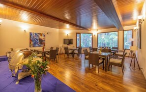 A dining area with wooden floors, a table, chairs, a TV, and windows with a forest view. - Bedzzz Xclusiv Baikunth Manali By Leisure Hotels (Manali)