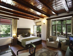 Une chambre avec un grand lit, un bureau, une chaise et un canapé. Elle est dotée de fenêtres avec des rideaux, d’un plafond en bois et offre une vue sur la verdure environnante.